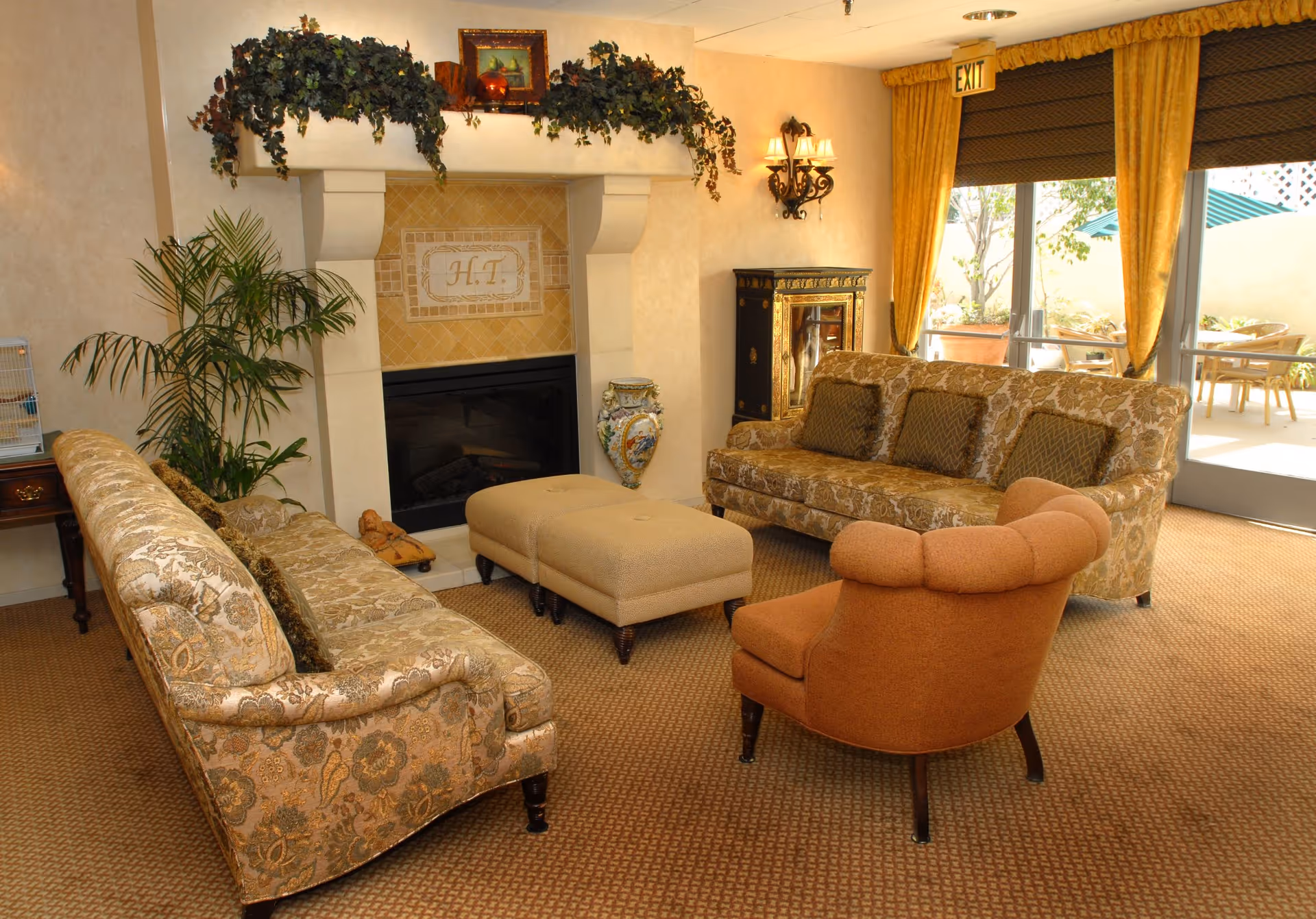 A cozy living room area in Harbor Terrace Retirement Community featuring two patterned sofas, an orange armchair, a beige ottoman, a fireplace with decorative greenery and framed art above it, a potted plant, a decorative vase, and large windows with yellow curtains leading to an outdoor patio with tables and chairs.