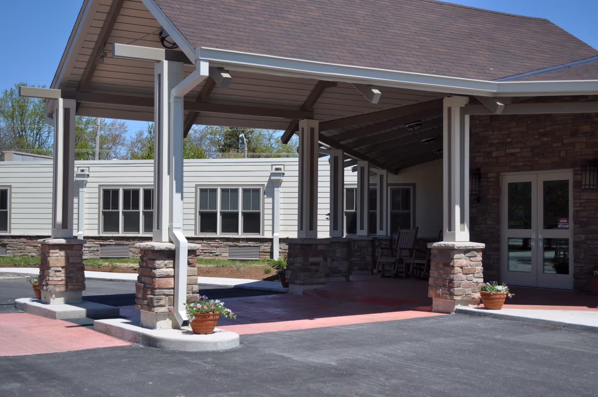 Covered entrance area of a senior living facility with stone pillars and a roof, potted flowers on the ground, and a double glass door entrance. The building has beige siding and multiple windows, with rocking chairs visible inside the covered area.