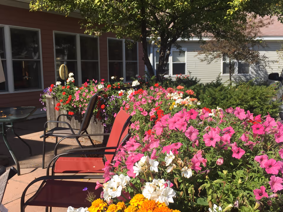 Outdoor patio with red chairs and abundant pink, white, and yellow flowers in planters beside a building.