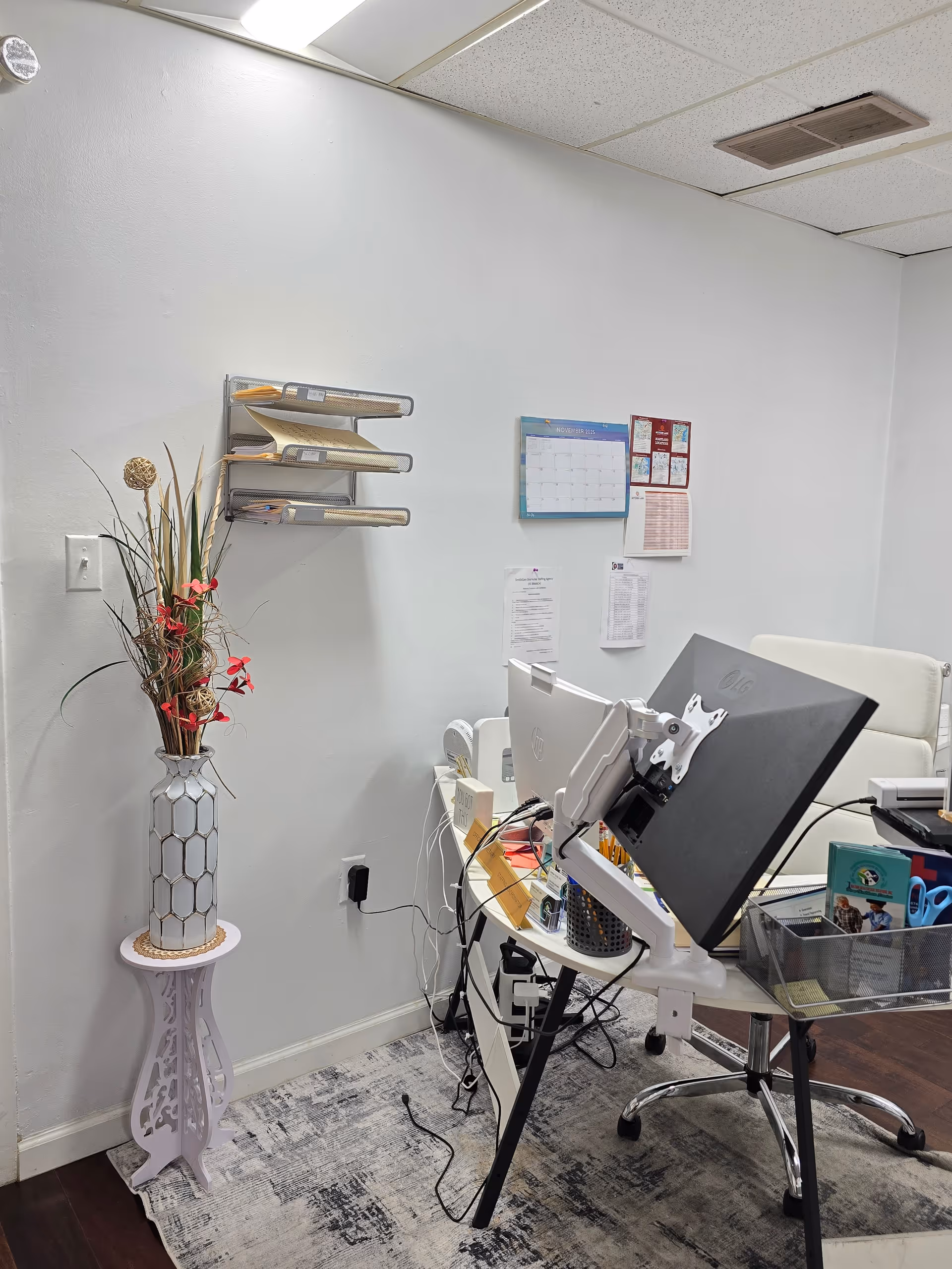 Office workspace with computer monitor, desk, calendar, and decorative vase against a light wall.