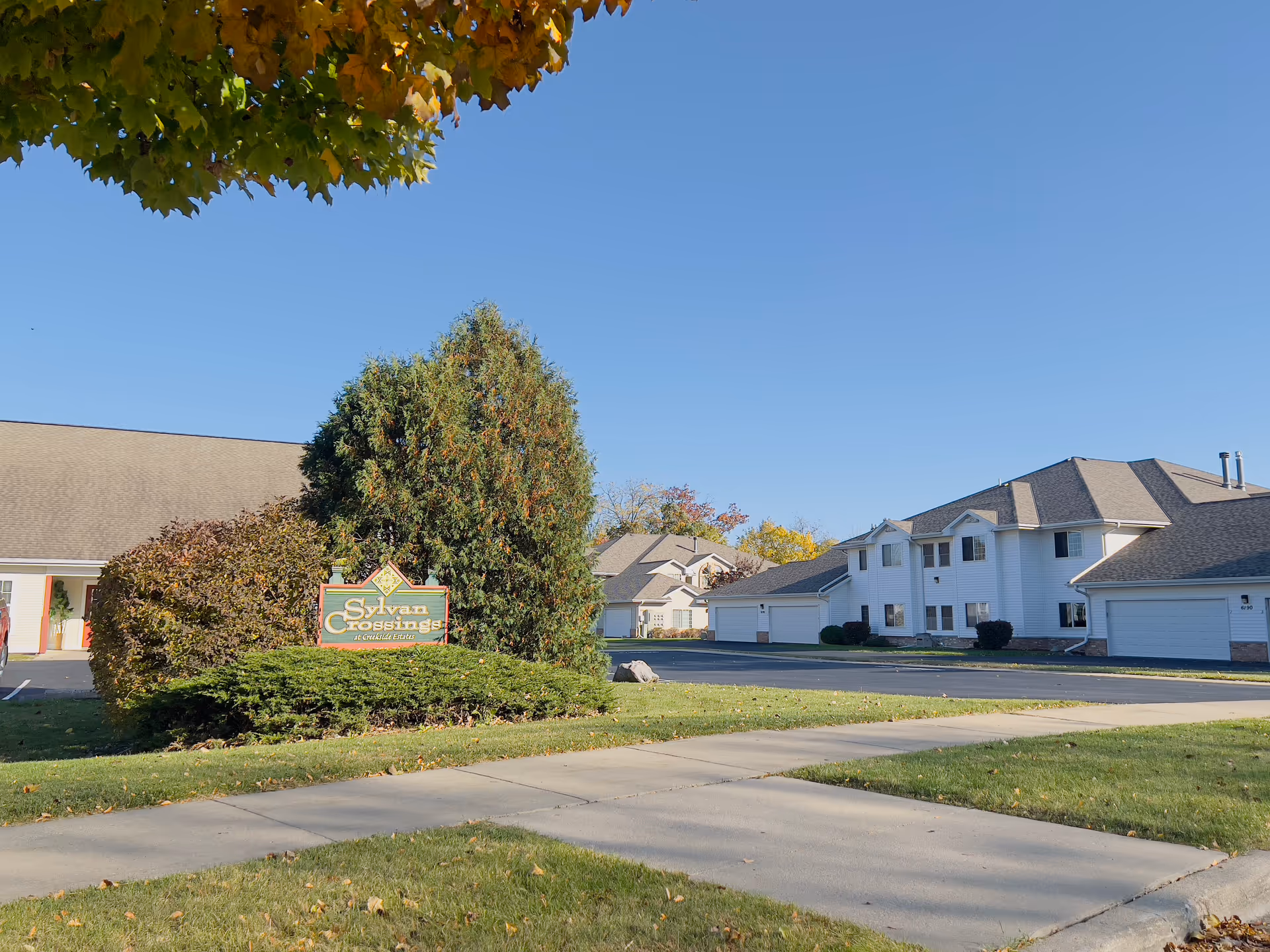 Exterior view of a senior living complex with white two-story buildings, a landscaped lawn, sidewalk, and a 'Sylvan Crossings' sign under a clear blue sky.