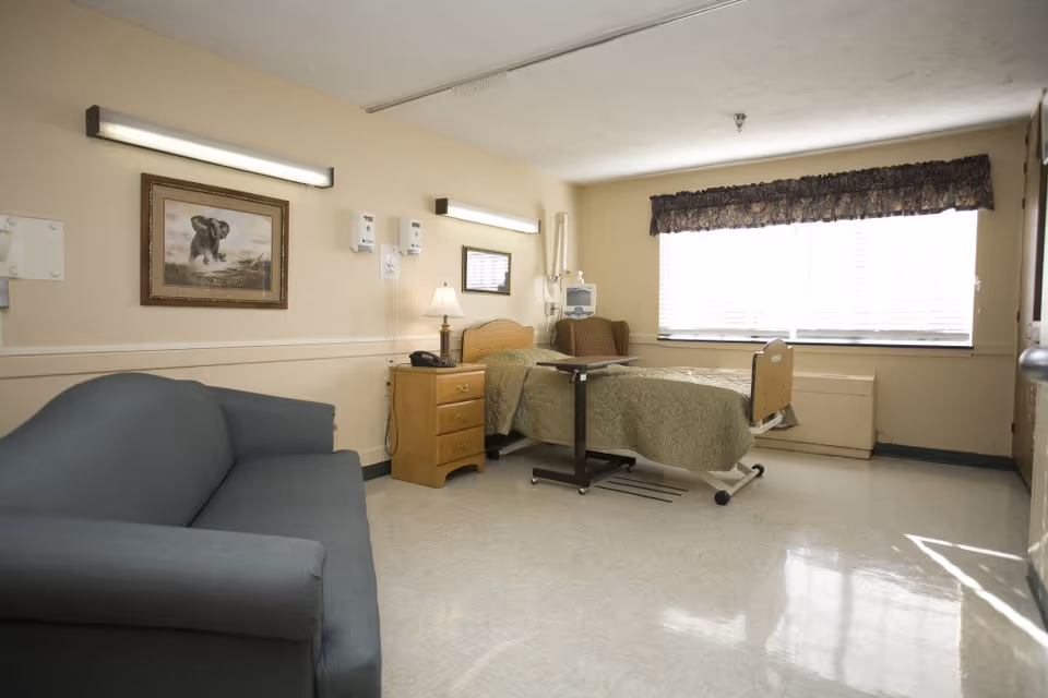 A hospital patient room with a bed, sofa, nightstand, lamp, and large window.