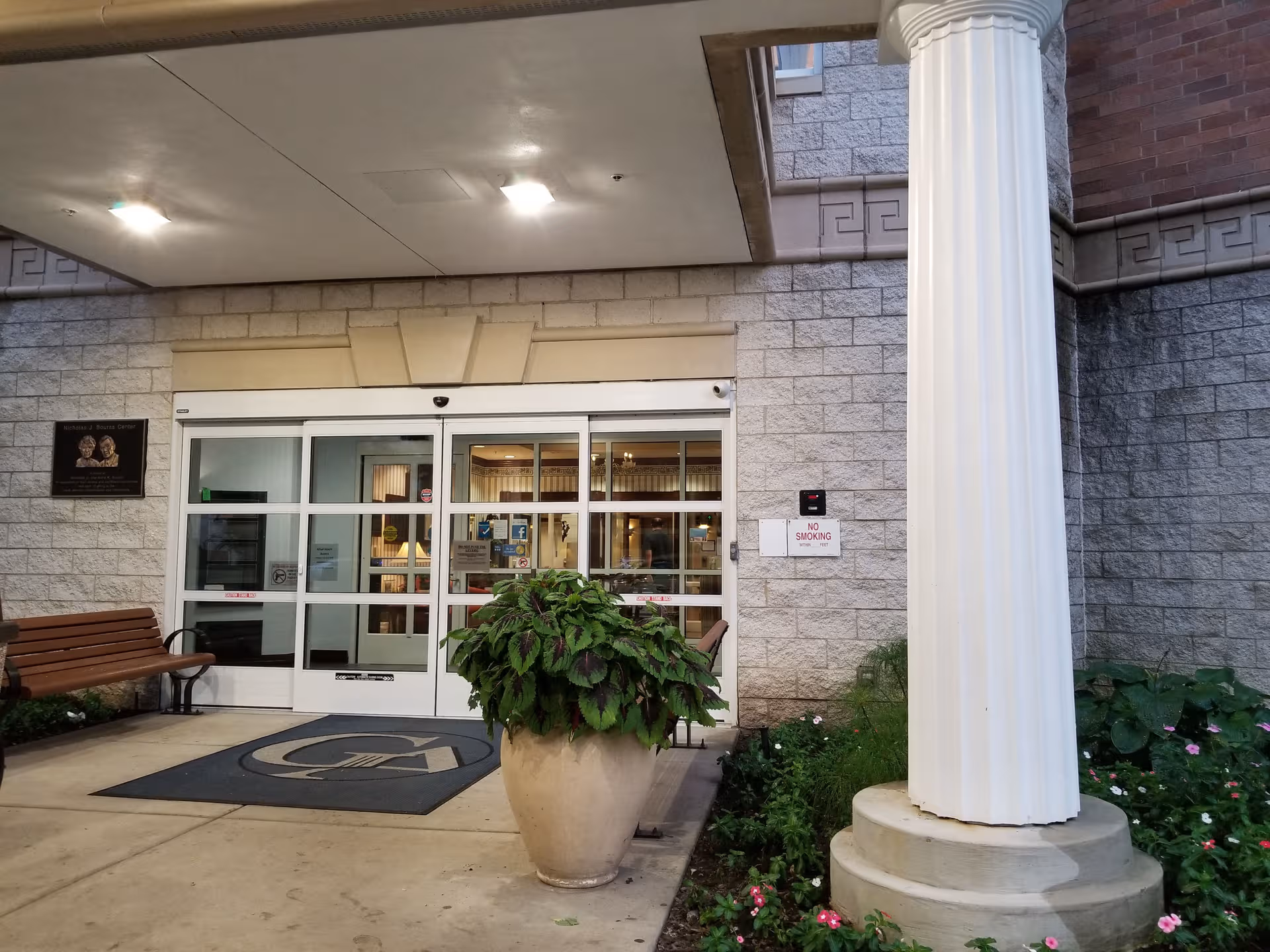 Entrance to Greek American Rehabilitation & Care Centre with glass doors, bench, column, and plants.