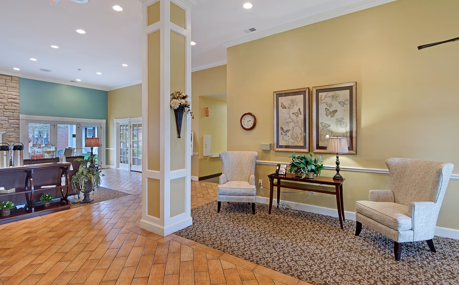 A senior living facility lobby area with two beige armchairs on a patterned carpet next to a wooden table with a lamp and a plant. The walls are painted yellow and decorated with two framed butterfly prints and a round clock. The floor is a combination of wood and tile, and there is a pillar in the center with a wall-mounted flower arrangement. In the background, there is a seating area with a lamp, plants, and large glass doors leading outside.
