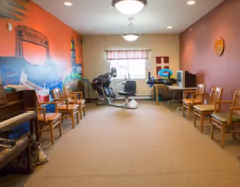 Sunlit interior activity room with chairs along the walls and a resident using equipment.