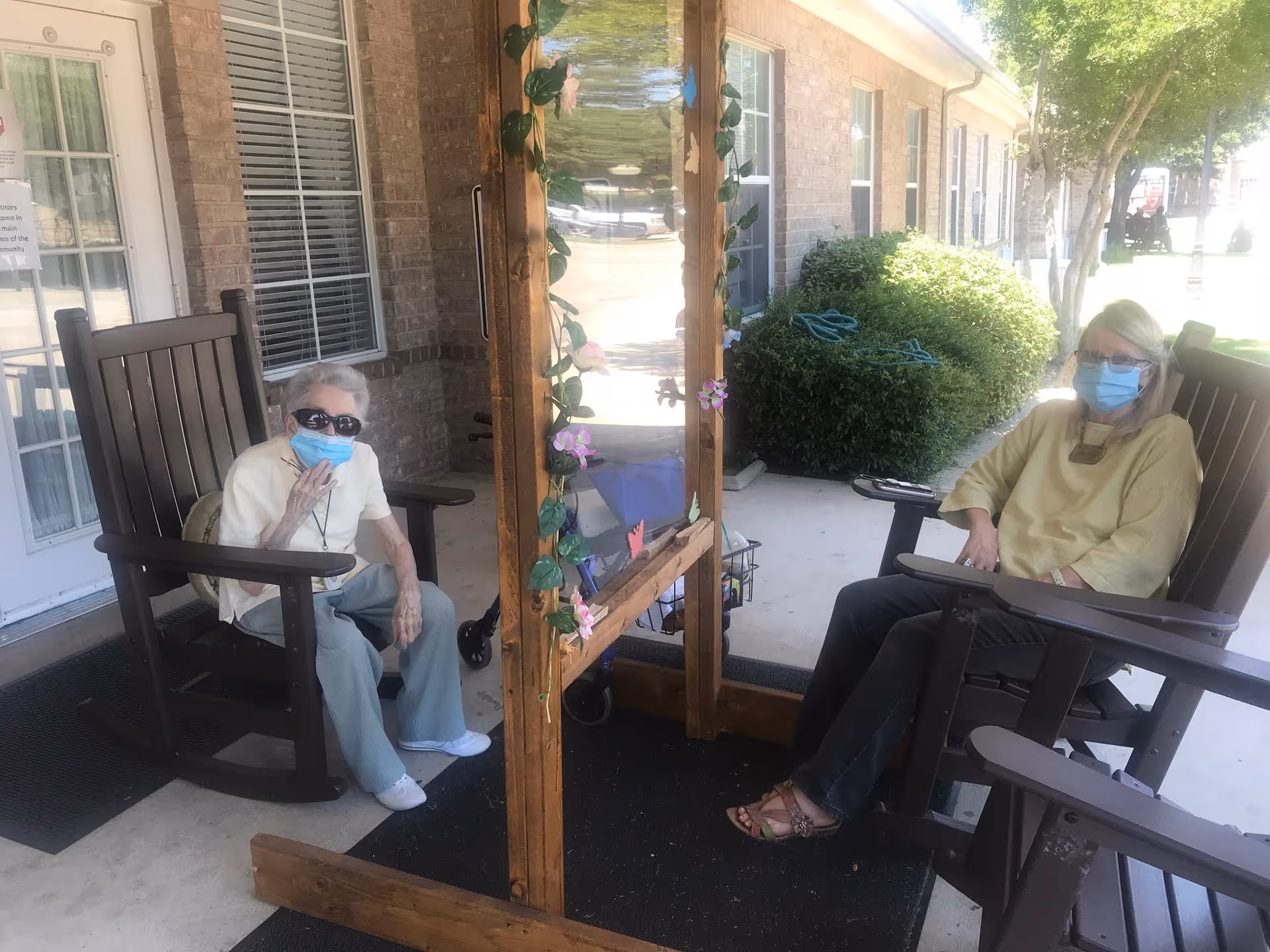 Two seniors wearing masks sit in rocking chairs on a porch outside a facility.