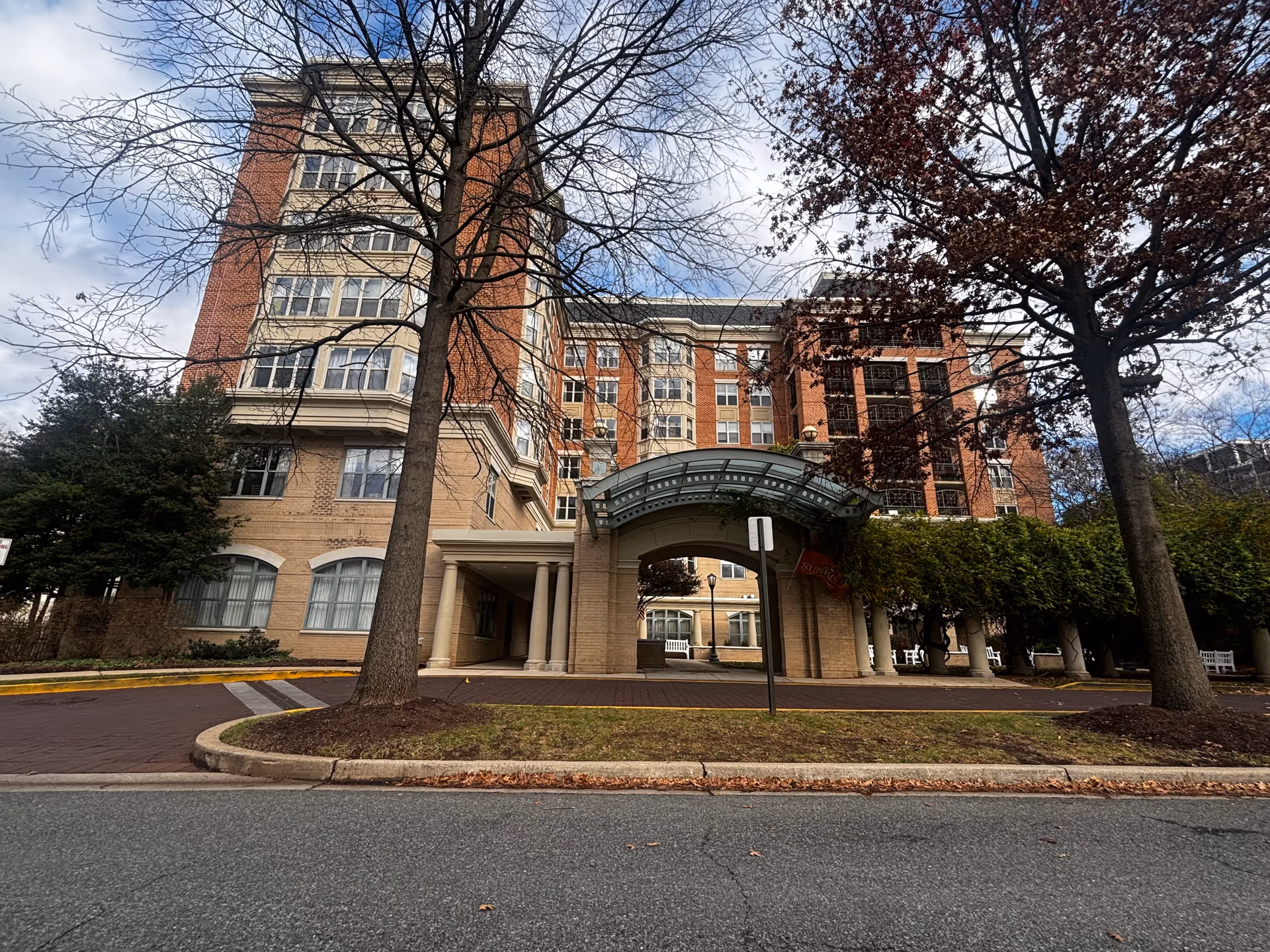 Exterior view of a multi-story brick and beige building with a covered entrance supported by columns. There are leafless trees and some greenery around the building, and a paved road in the foreground.