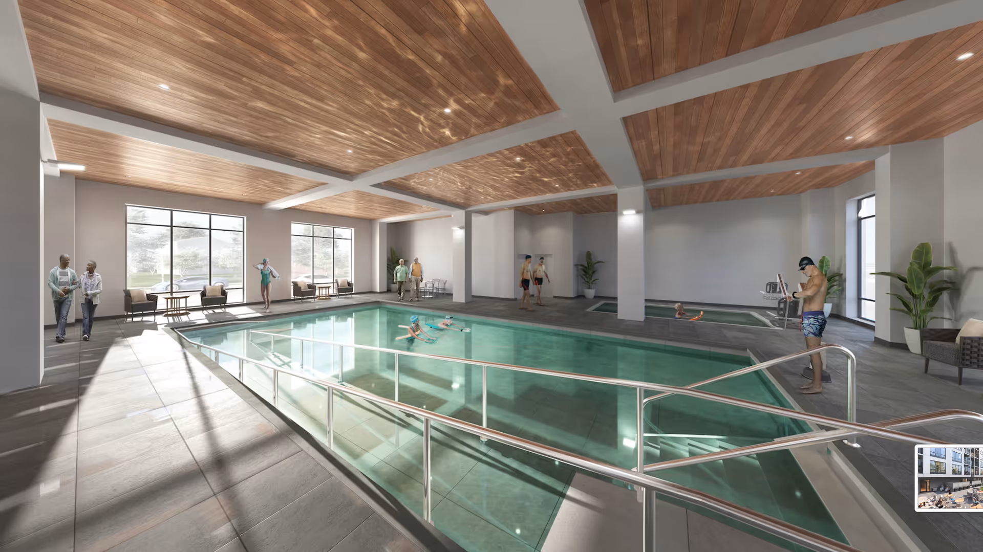 Indoor swimming pool area with a wooden ceiling and large windows letting in natural light. Several people are present, including two swimmers in the pool, a man standing by the poolside in swimwear, and others walking or standing nearby. There are chairs and tables along the windows and potted plants in the corners.