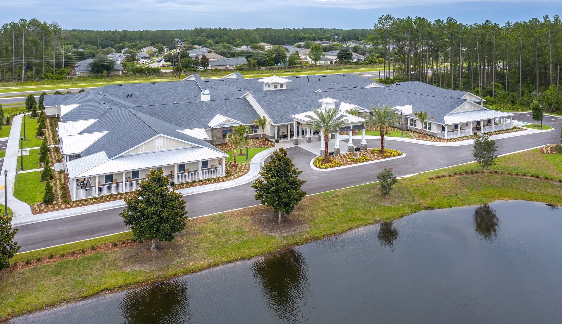 Aerial view of The Addison of St. Johns senior living facility showing a large, single-story building with a gray roof and white exterior. The building is surrounded by landscaped grounds with palm trees, a driveway, and a small pond in the foreground. There are residential homes and trees in the background.