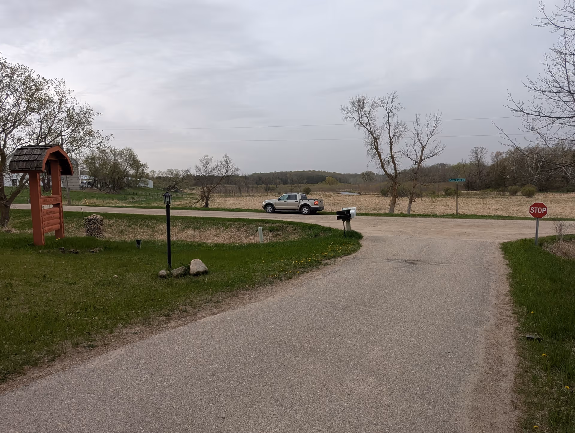 Rural entrance with driveway, stop sign, mailbox, and red wooden shelter beside bare trees.