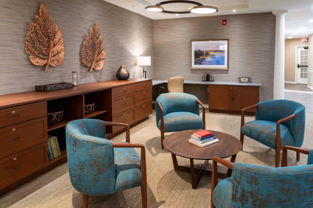 A cozy seating area in an assisted living facility featuring four teal upholstered armchairs arranged around a round wooden coffee table with a stack of books. The room has wooden cabinets along the walls, decorative leaf wall art, a table lamp, and a framed landscape painting. The space is well-lit with ceiling lights and has a neutral-toned carpet and wallpaper.
