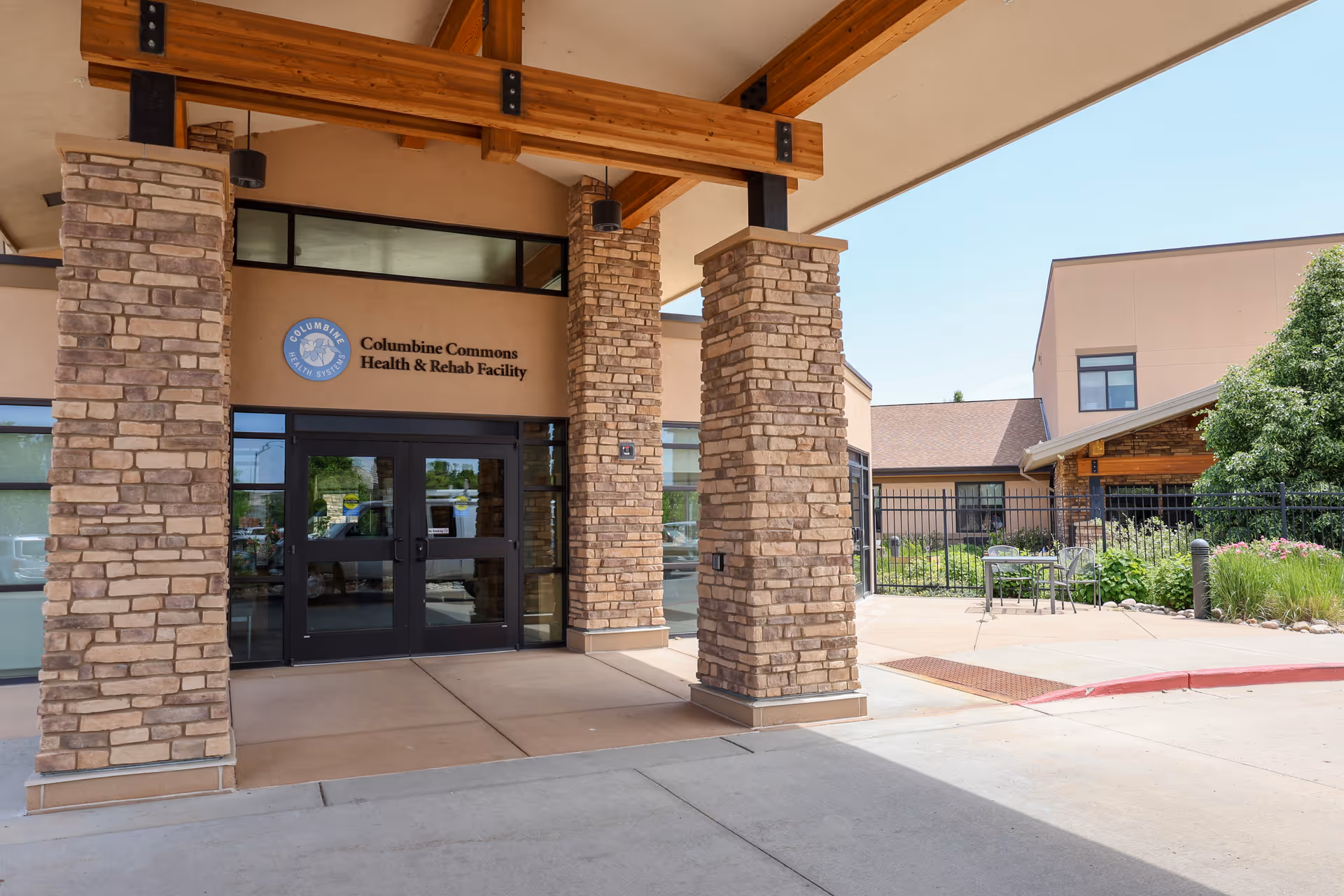 Entrance of Columbine Commons Health & Rehab Facility with stone pillars supporting a wooden beam roof overhang, glass double doors, and a small outdoor seating area with a table and chairs surrounded by greenery.