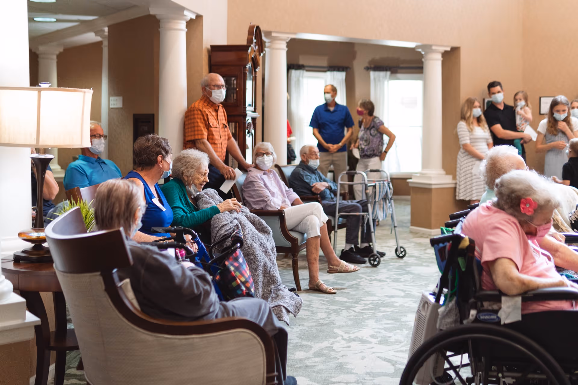 A group of elderly residents and visitors wearing face masks are gathered in a spacious, well-lit common area with beige walls and white columns. Some residents are seated in wheelchairs and chairs, while others stand or use walkers. The atmosphere appears calm and social.