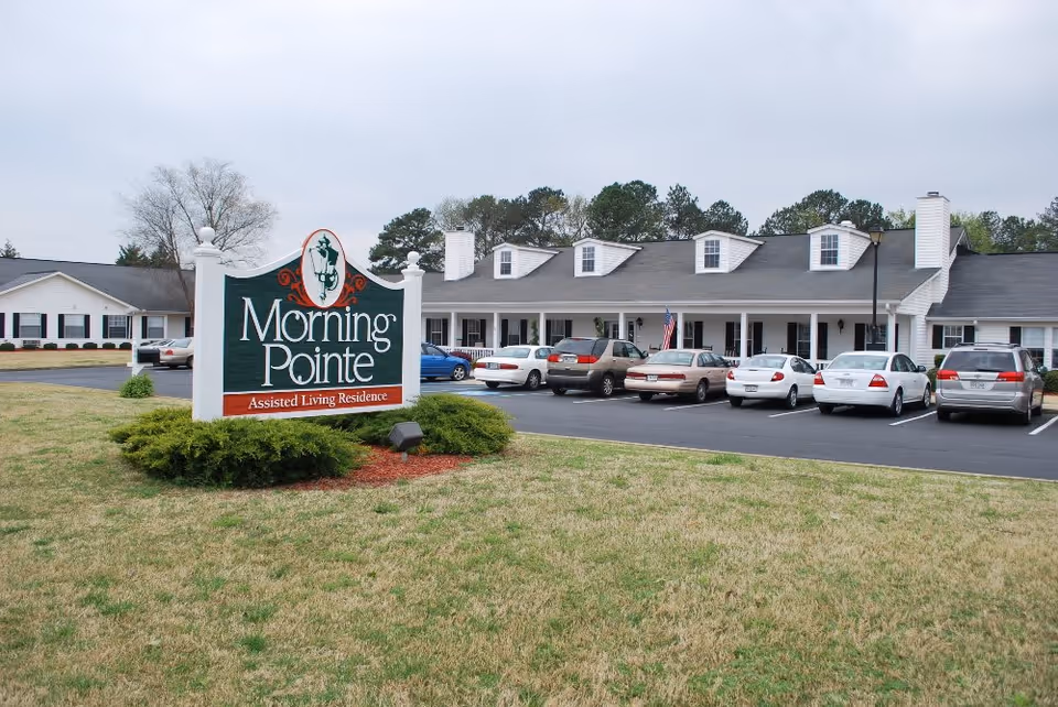 Exterior view of Morning Pointe Assisted Living Residence in Calhoun, showing a large sign in the foreground and a white building with multiple dormer windows and parked cars in front.