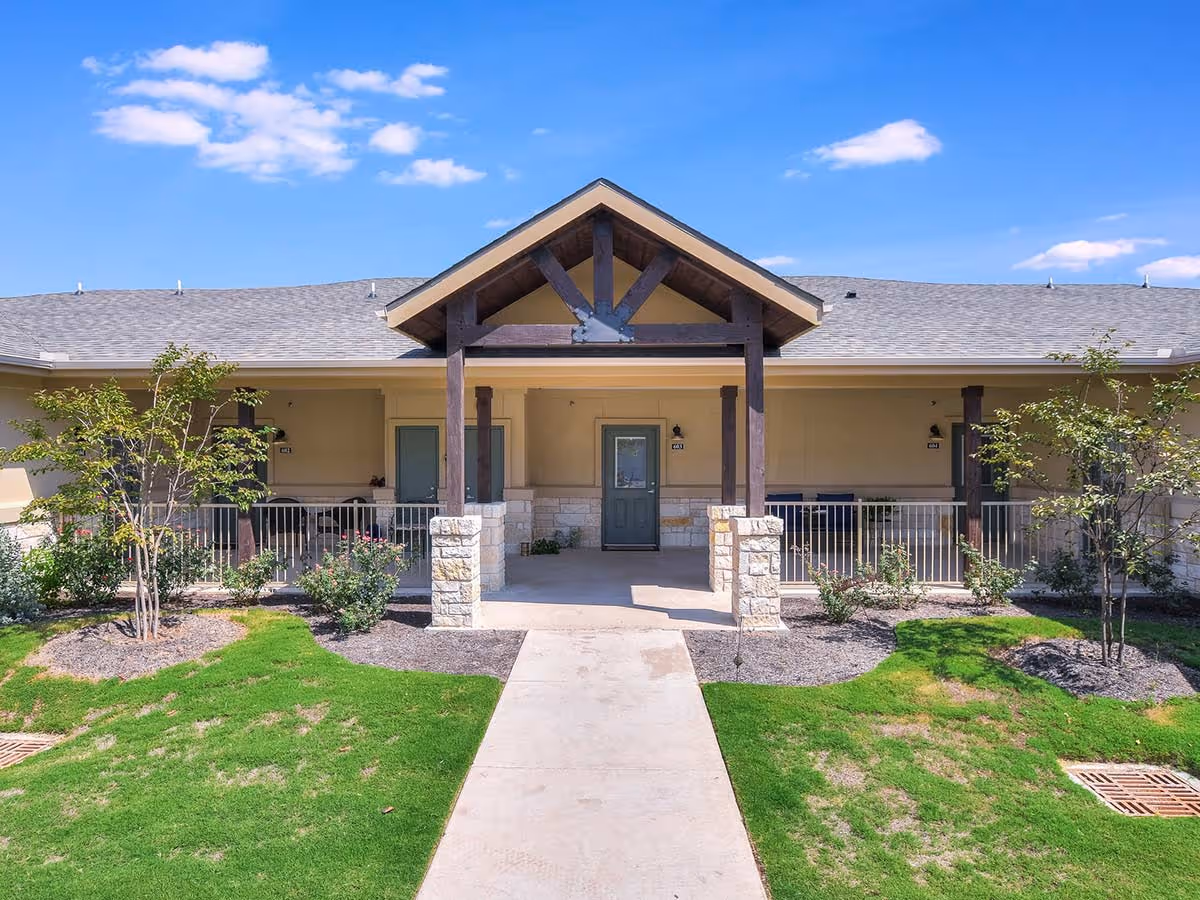 Front exterior view of a single-story senior living facility building with a covered entrance supported by wooden beams and stone pillars. The building has a gray shingled roof, beige walls, green doors, and a well-maintained lawn with small trees and shrubs under a blue sky with scattered clouds.