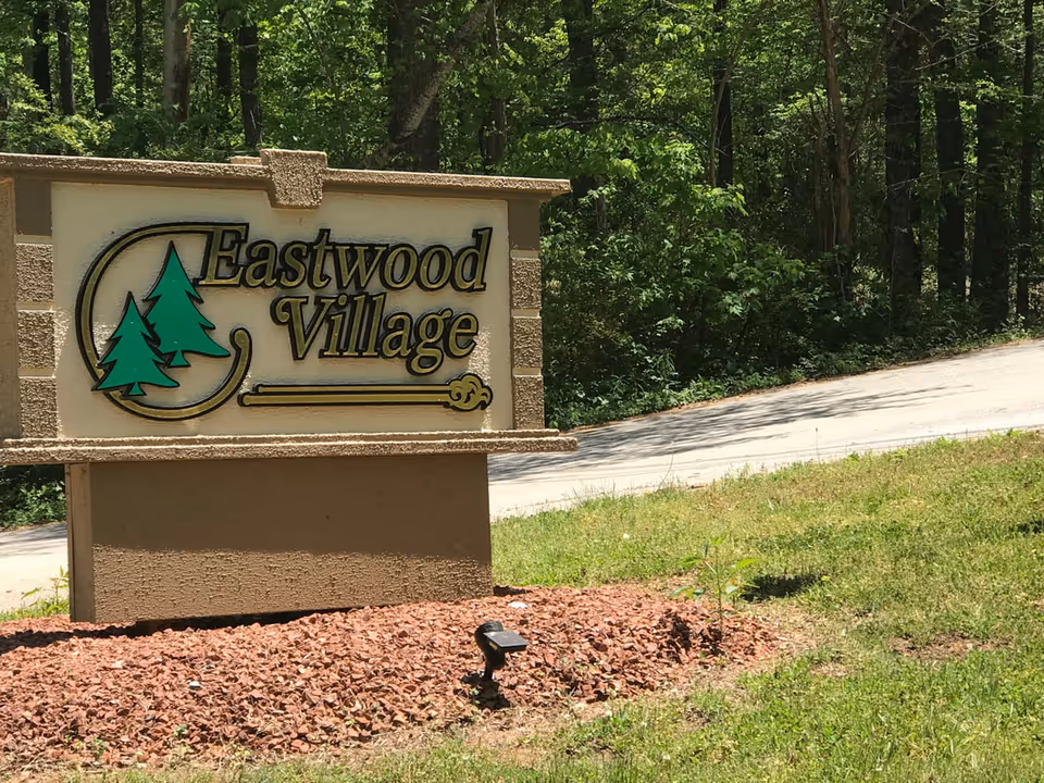 Stone entrance sign reading Eastwood Village beside a wooded road and trees.
