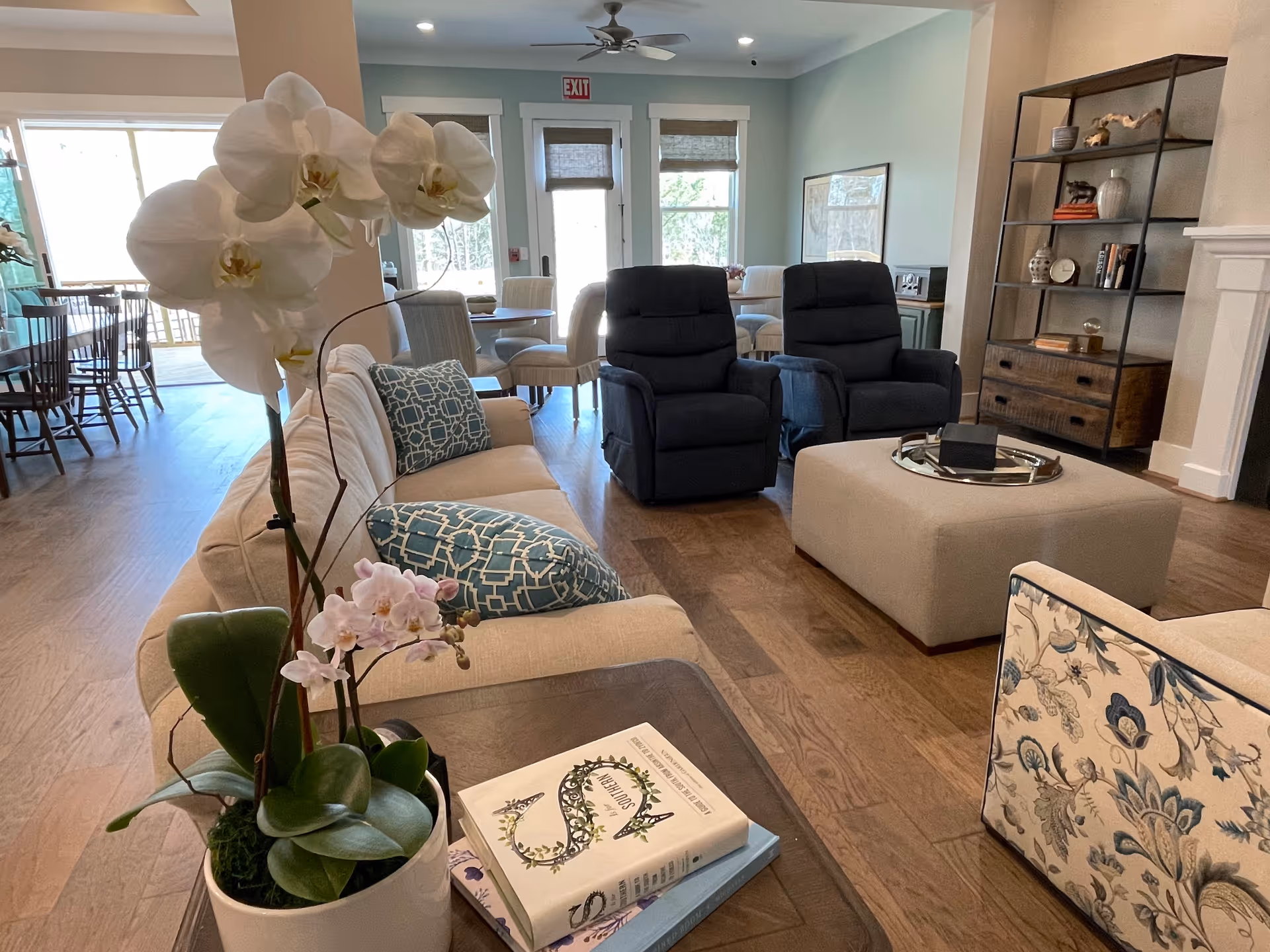Cozy senior living lounge with beige sofa, blue recliners, orchid plant, and coffee table.
