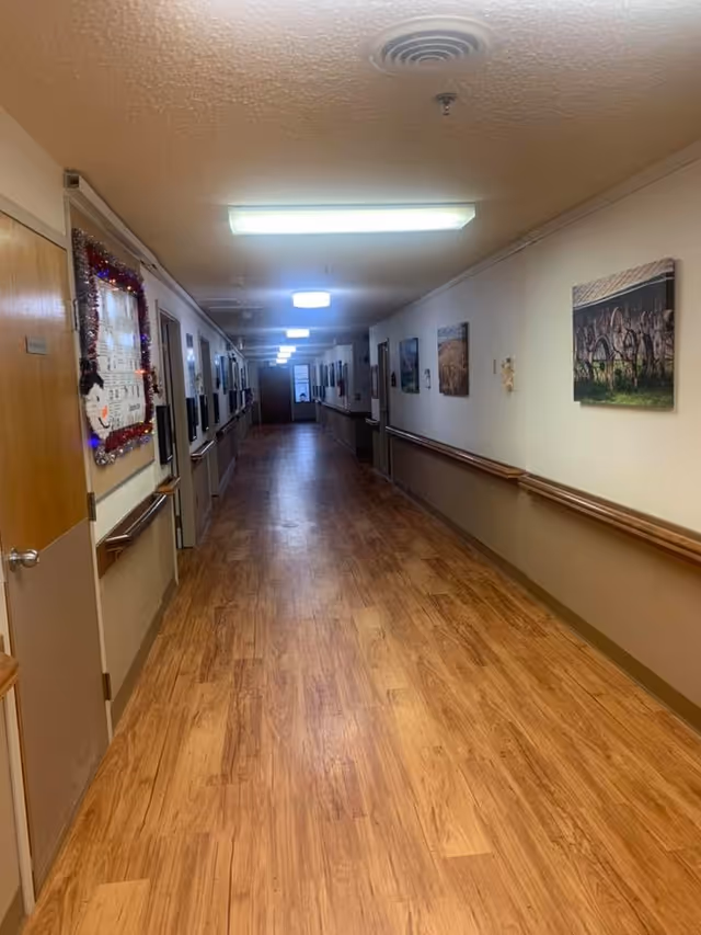 Long, well-lit interior hallway with wood-look flooring, handrails, doors and framed artwork along the walls.