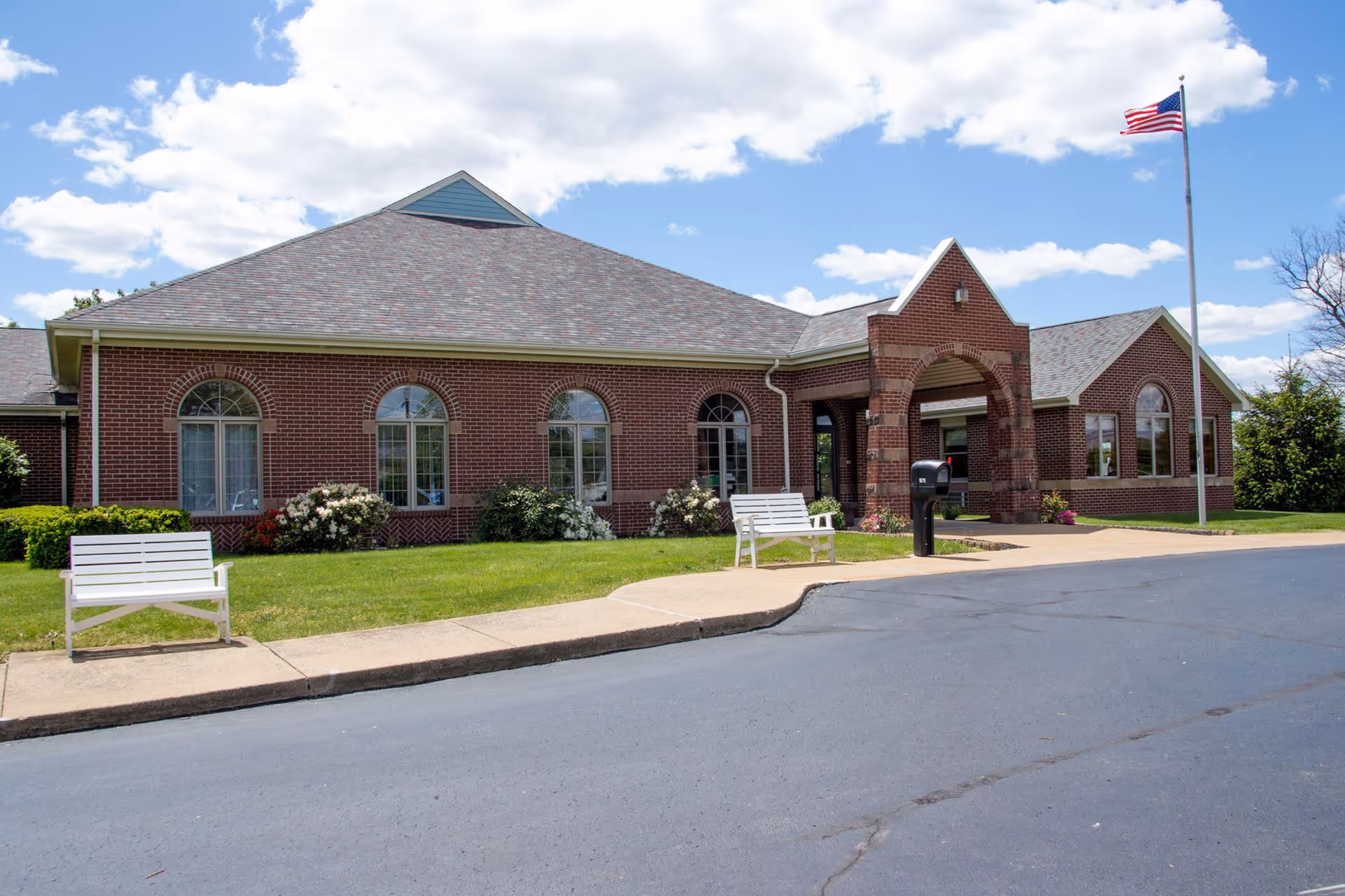 Single-story brick senior living building with arched windows, white benches on the lawn, an entrance archway, and an American flag on a pole.