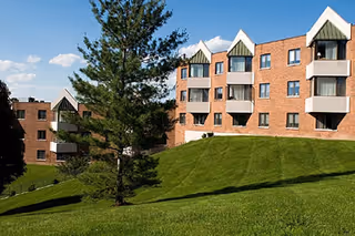 Brick senior living building with balconies on a grassy hill and trees.