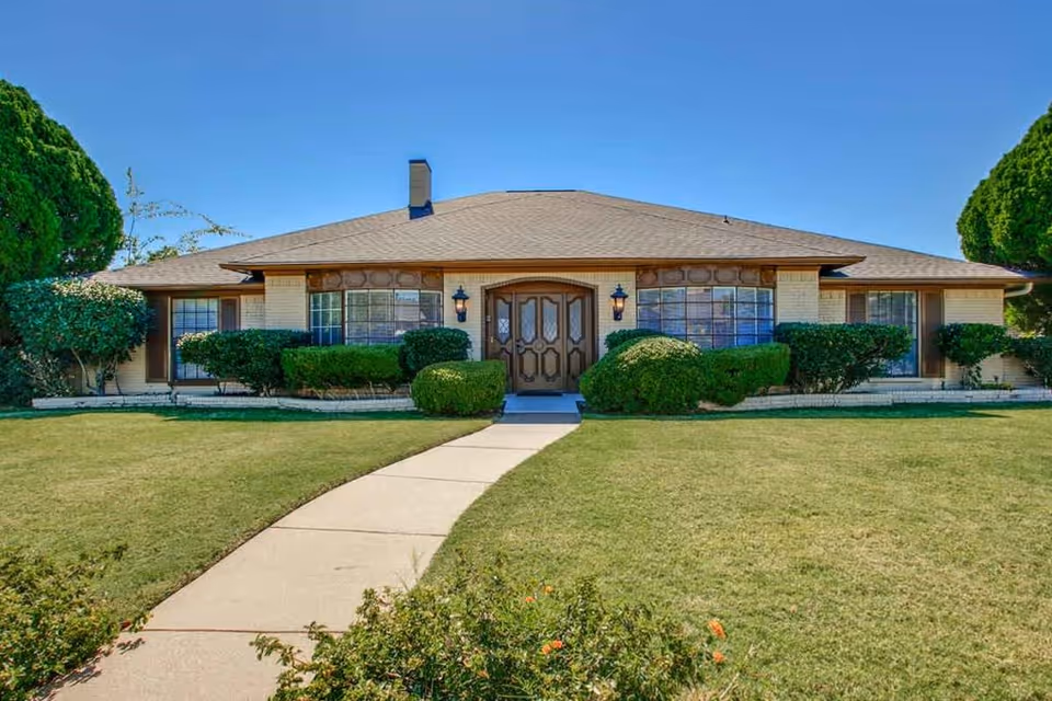 Front exterior view of a single-story brick building with a central entrance door flanked by two large windows and surrounded by neatly trimmed bushes and a well-maintained lawn under a clear blue sky.
