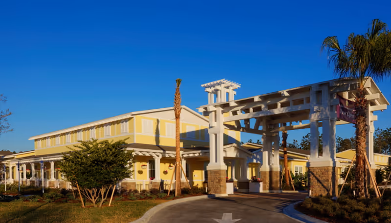 Front entrance and porte-cochere of a yellow senior living building with palm trees and a curved driveway.