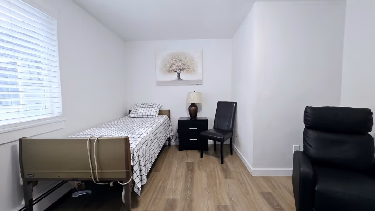 A small, clean senior care bedroom with a bed, nightstand, lamp, and recliner.