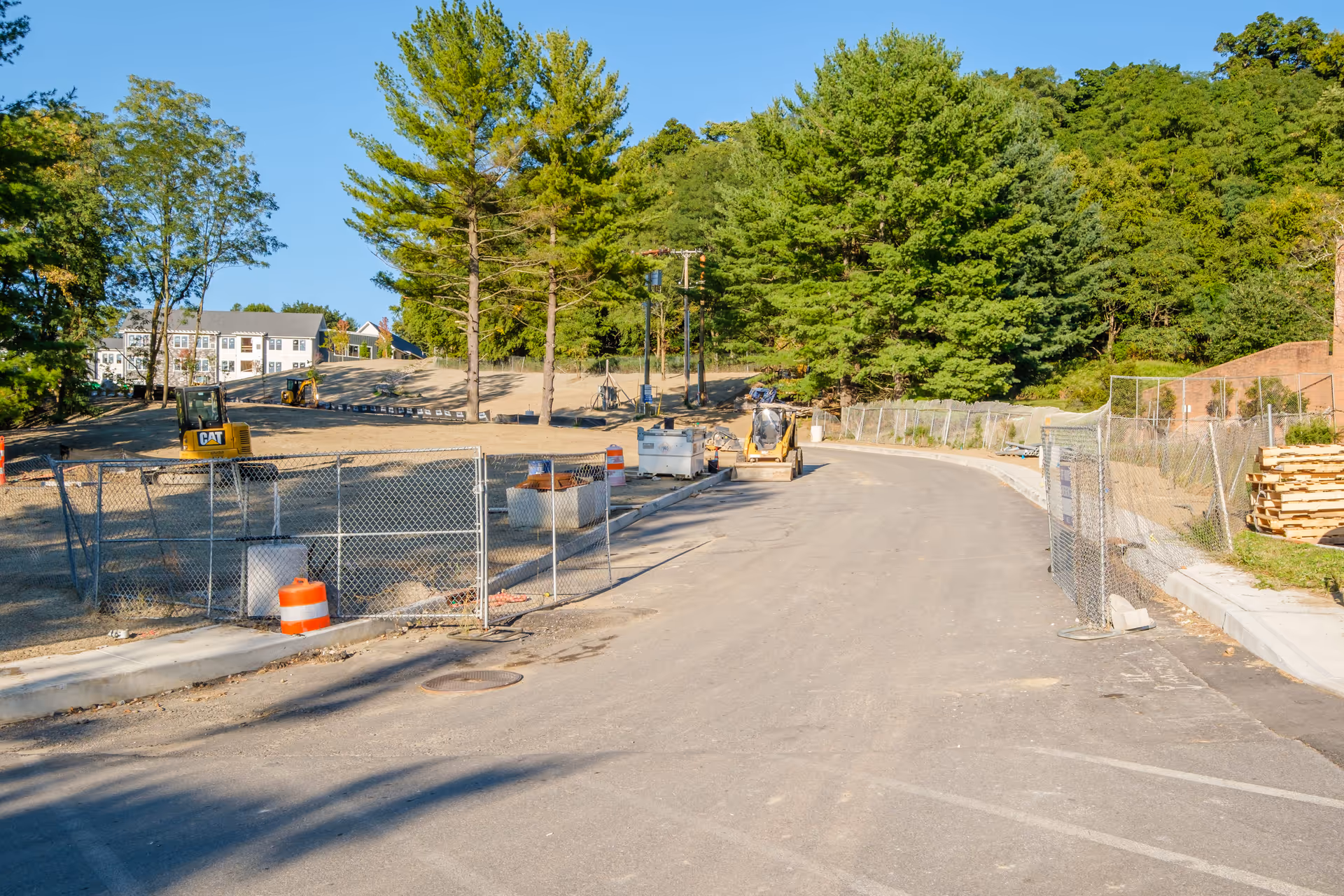 A construction site with a paved road curving to the right, surrounded by temporary chain-link fencing and orange traffic barrels. There are construction vehicles and equipment on the dirt area to the left, with trees and a building in the background under a clear blue sky.