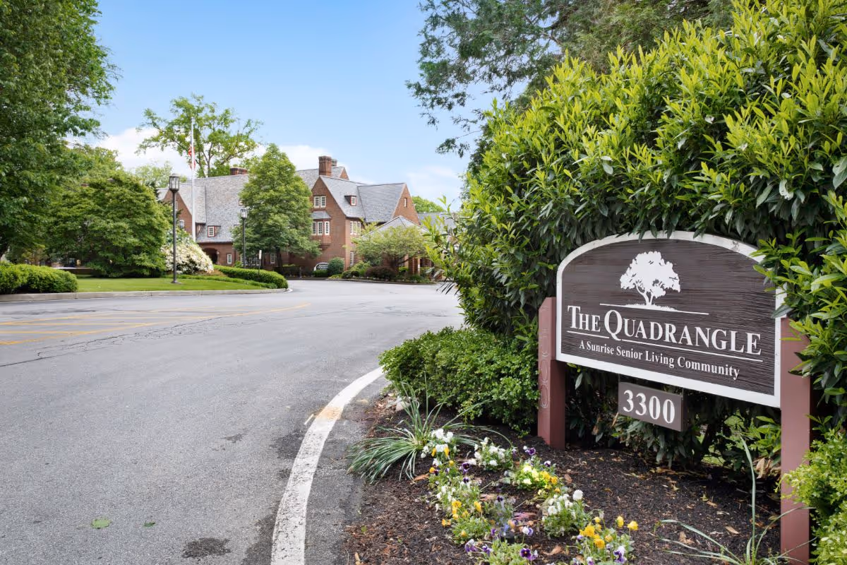 Entrance sign for The Quadrangle senior living community next to a driveway and landscaped beds with the brick residence visible in the background.