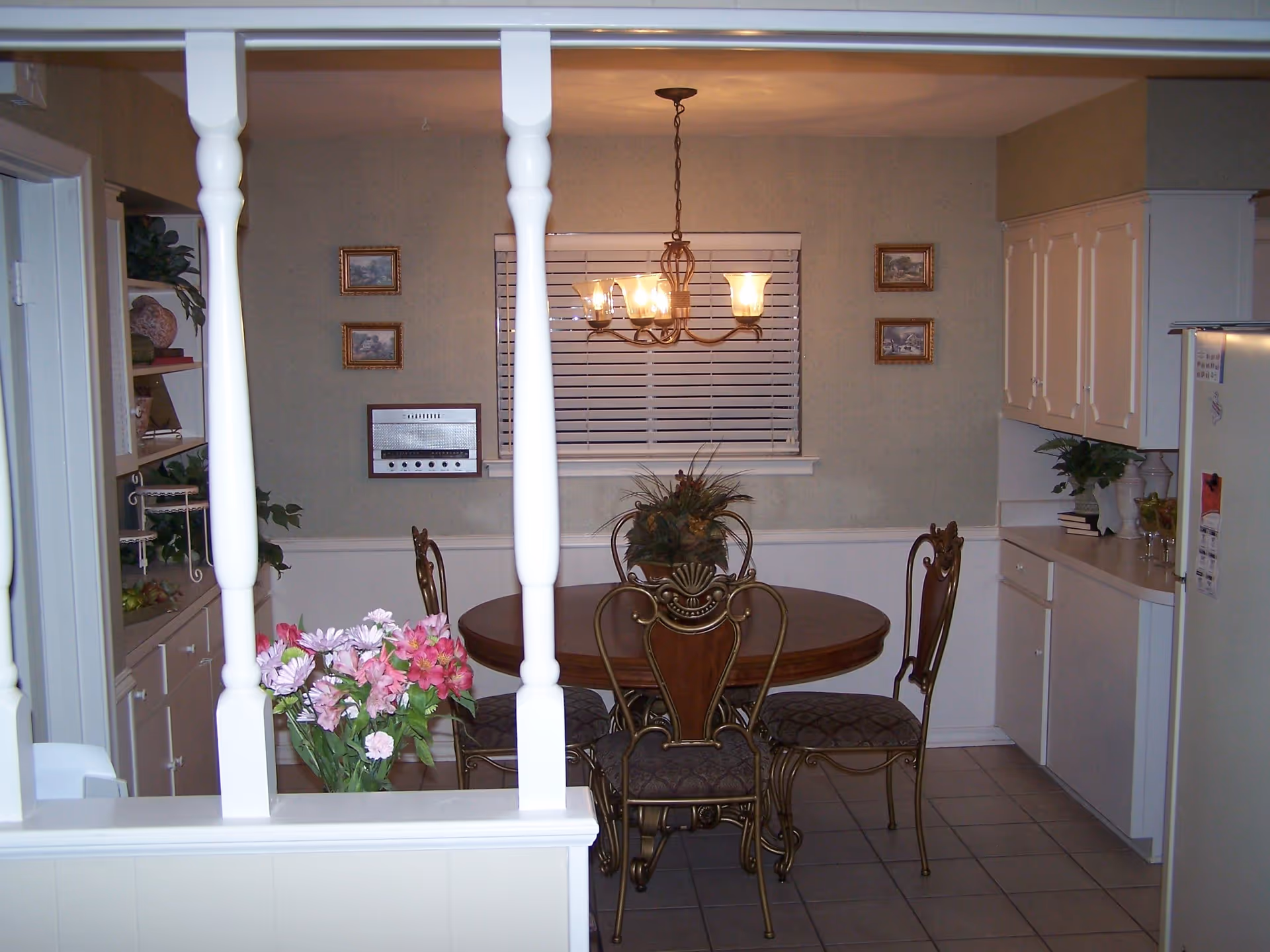 Interior view of a dining area with a round wooden table and four ornate chairs with cushioned seats. A chandelier with five lights hangs above the table. The room has light-colored walls with four small framed pictures and a window with closed blinds. To the right, there are white kitchen cabinets and a refrigerator. To the left, there are shelves with decorative items and plants. A vase with pink and white flowers is visible in the foreground.