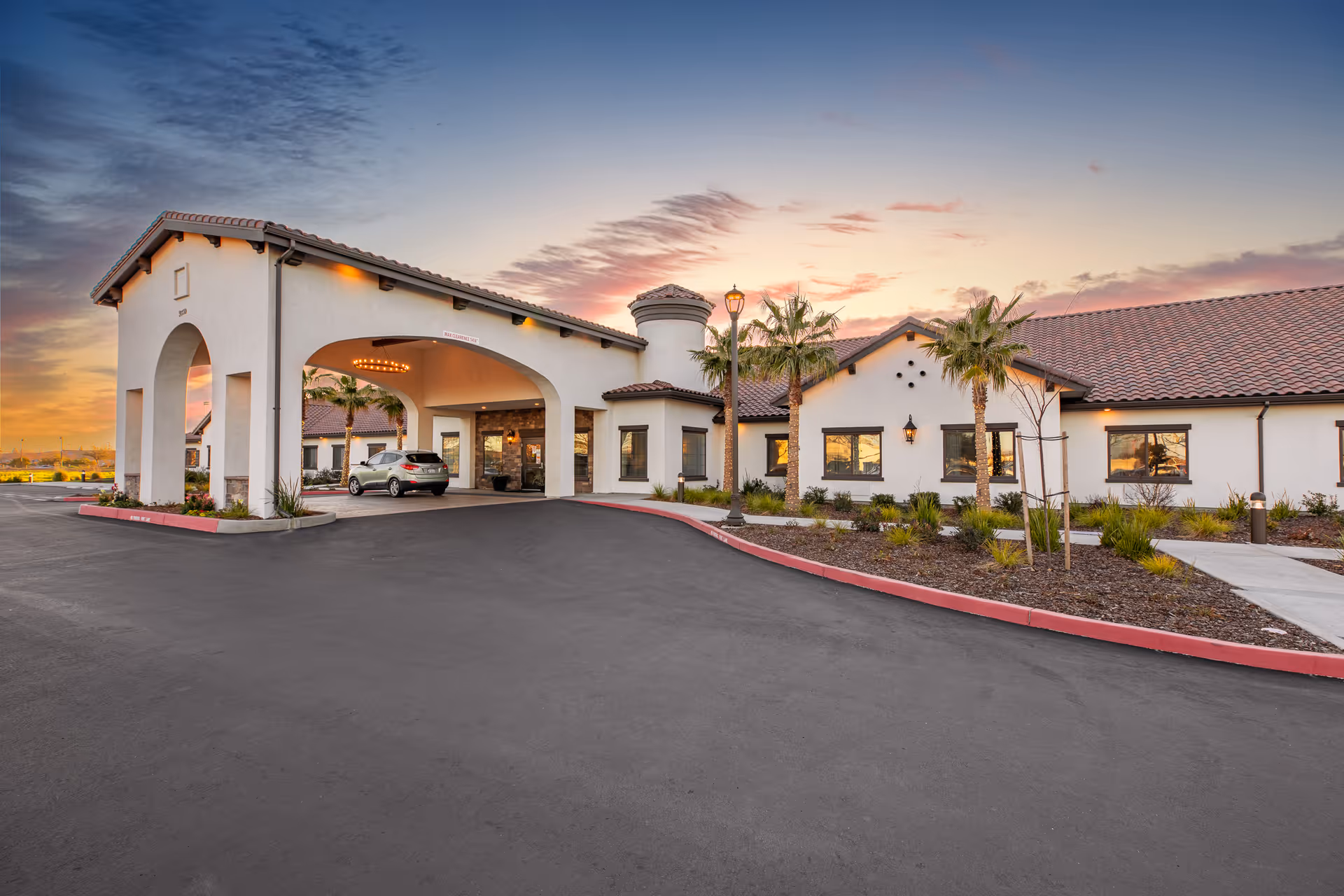 Exterior view of Gardens at Laguna Springs entrance with arches, palm trees, and sunset sky