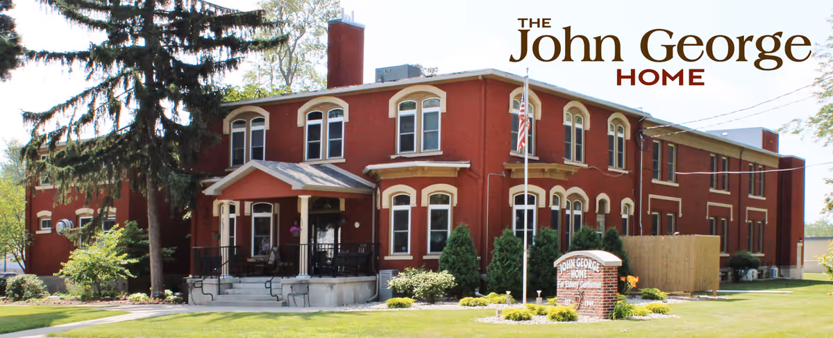 Two-story red brick senior home with a front porch, flagpole, landscaped lawn and a sign reading 'John George Home'.