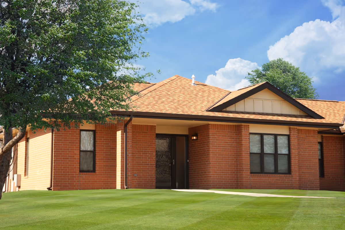 Single-story red brick building front with a covered entrance, manicured green lawn, and a tree under a blue sky.