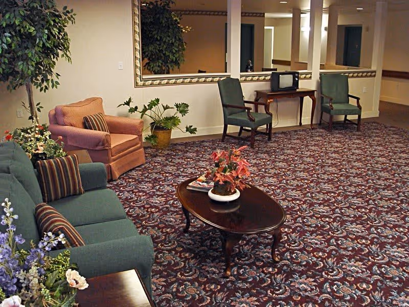 A senior living facility common area with patterned carpet, a green sofa with striped pillows, a pink armchair, two green armchairs, a wooden coffee table with a potted plant and magazines, and a large mirror on the wall. There are also several potted plants around the room.