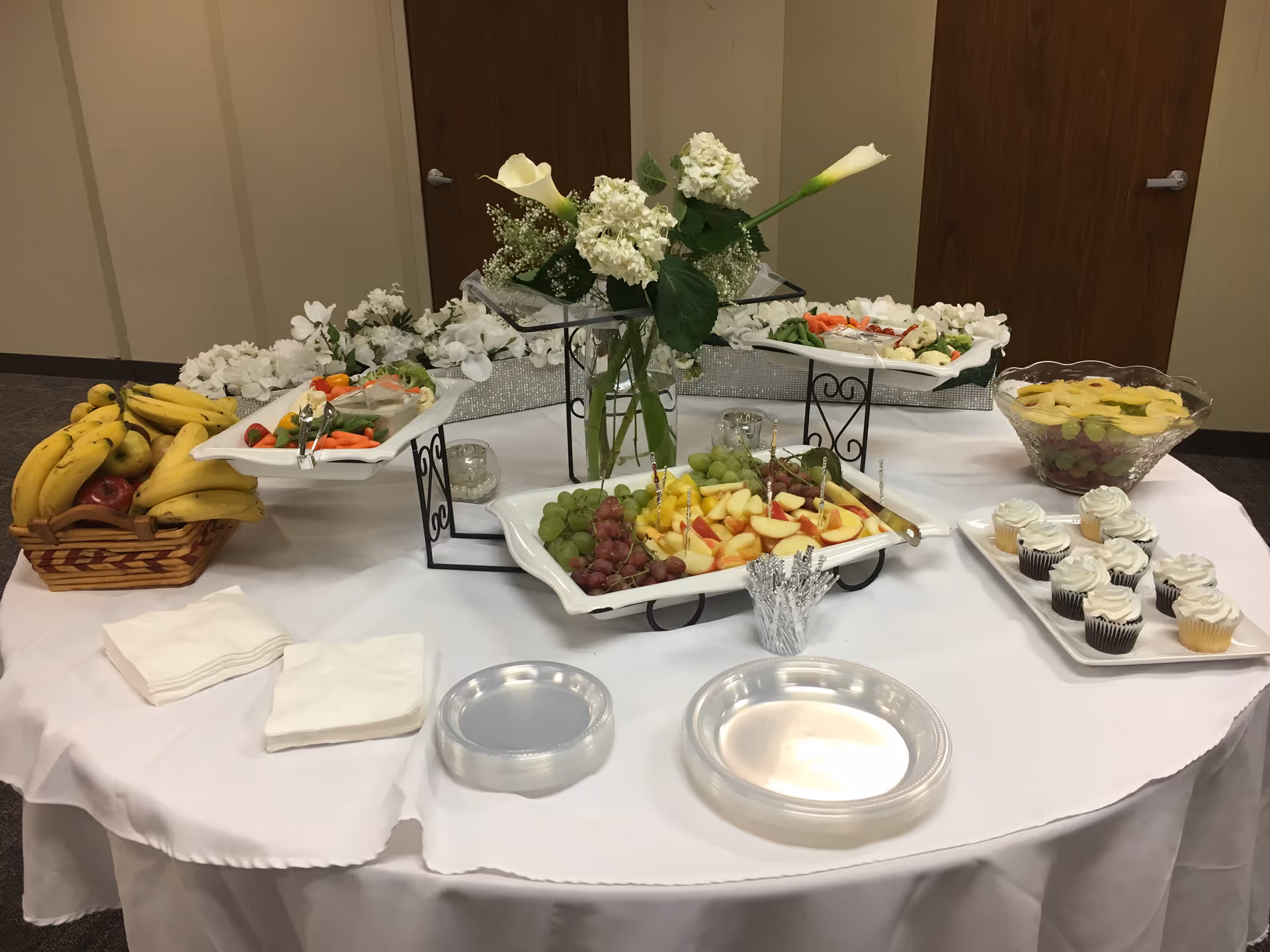 Buffet table with fruit, cupcakes, and flowers in a dining room.