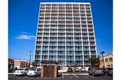 Tall, multi-story building with many balconies and a parking lot in front.