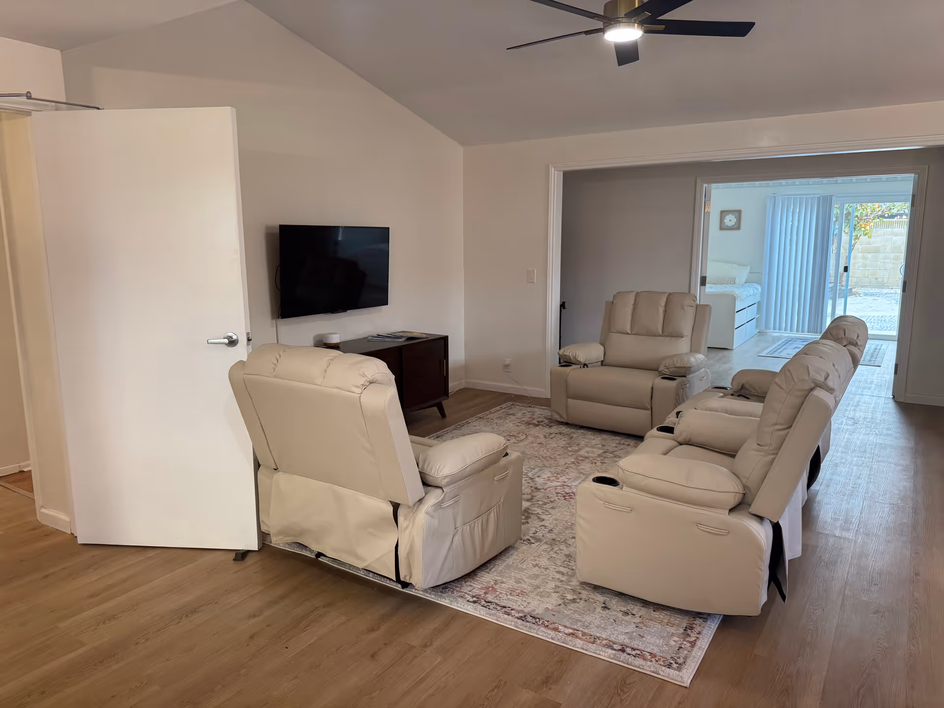 Living room with beige recliners, a rug, wall-mounted TV, ceiling fan, and glass door to outside.