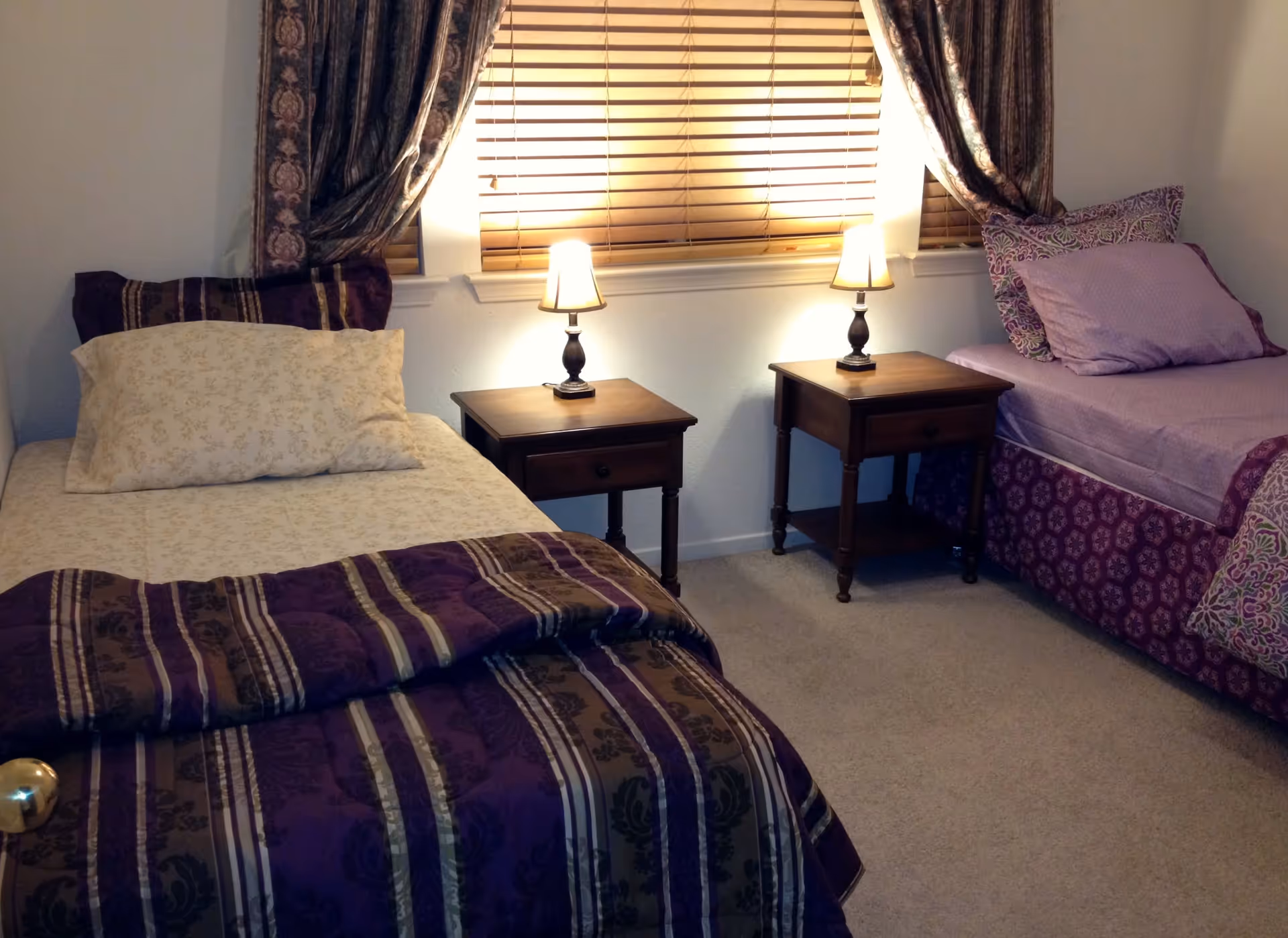 Two twin beds separated by wooden nightstands with lamps under a window with blinds and curtains.