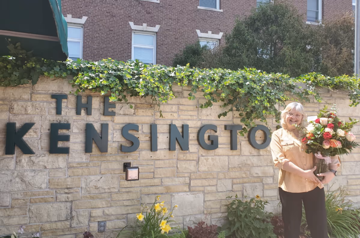 Woman holds bouquet beside The Kensington sign on a stone wall outdoors.