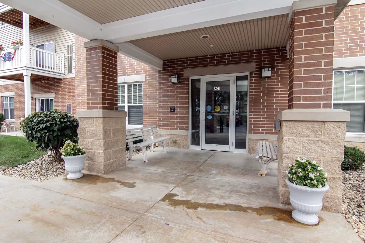 Brick building entrance with a covered porch, benches, and potted plants.