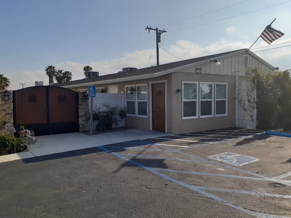 Exterior view of a single-story building with beige walls and a brown door, adjacent to a large wooden gate. There is a handicapped parking space in front, marked with blue lines and a sign. An American flag is flying on a pole attached to the building. Palm trees and a clear sky are visible in the background.