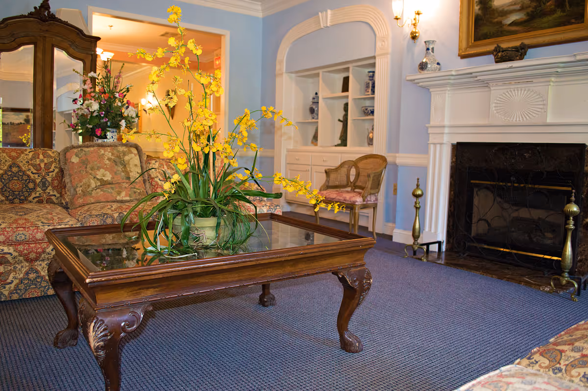 A cozy living room with a patterned sofa and floral cushions, a wooden coffee table with a glass top holding a large yellow orchid plant, a fireplace with decorative brass andirons, a built-in white shelving unit with blue and white vases, a wooden chair with a pink cushion, and a large floral arrangement in the background.