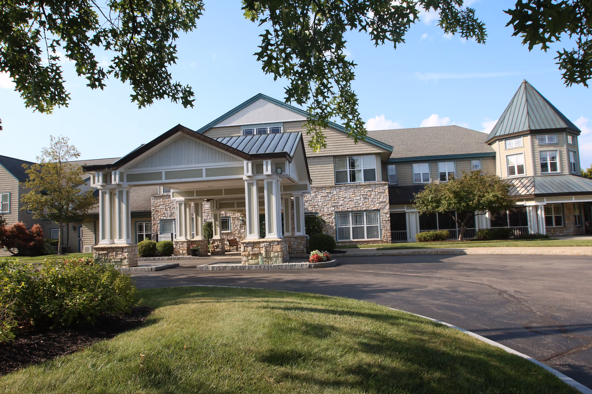 Exterior view of a senior living facility named Glen Eddy, featuring a large covered entrance with stone pillars, a paved driveway, well-maintained green lawns, and trees under a clear blue sky.