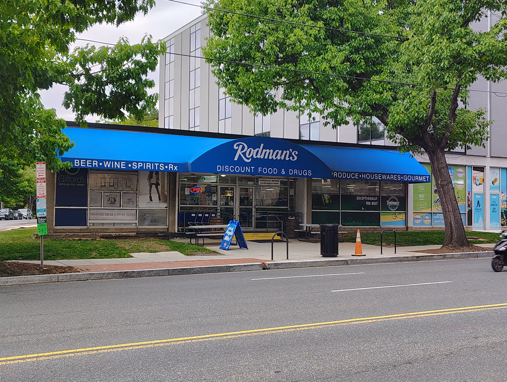 Rodman's storefront with blue awning across a single-story building on a city street, trees lining sidewalk.