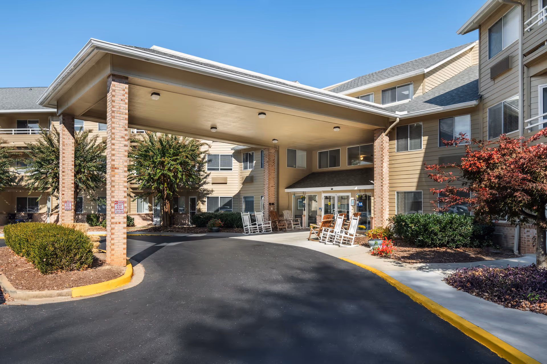 Entrance of Haywood Estates by Barclay House featuring a covered driveway with brick pillars, several white and wooden rocking chairs on the sidewalk, landscaped bushes and trees, and a clear blue sky.