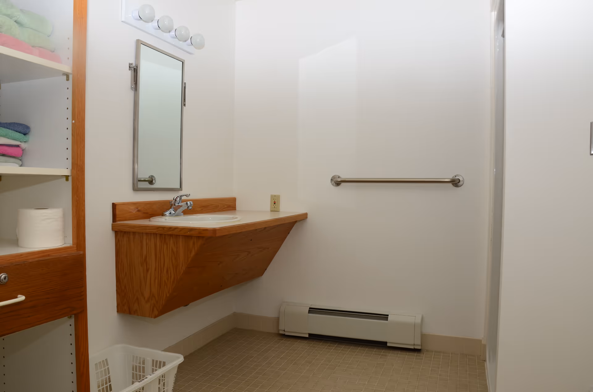 A bathroom with a wall-mounted wooden sink vanity, a rectangular mirror above the sink, a towel bar on the wall, and a shelf unit with folded towels and a roll of toilet paper. The floor is tiled and there is a baseboard heater below the towel bar.