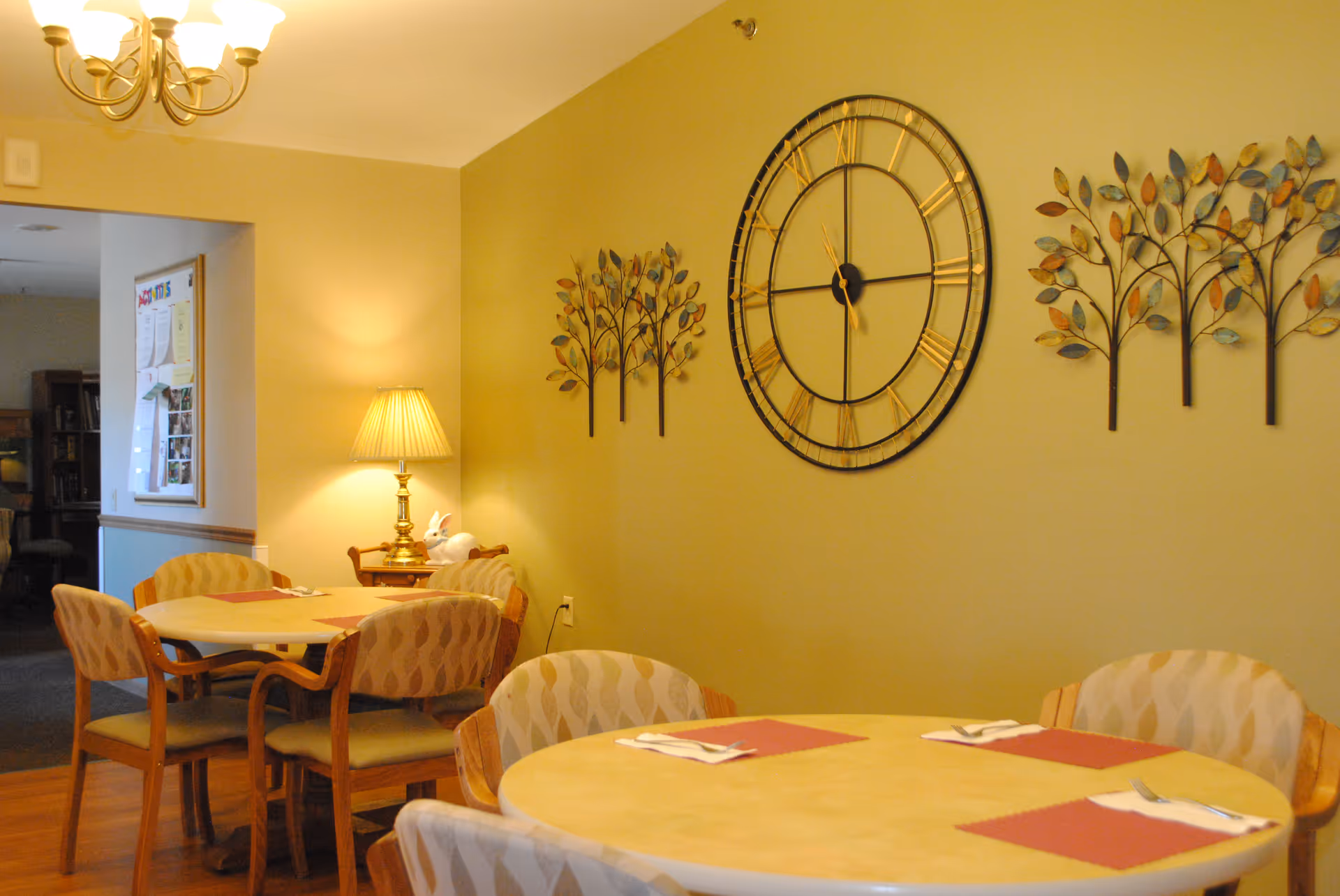 A cozy dining area with round tables and cushioned chairs. The walls are painted a warm yellow and decorated with a large round clock and metal tree wall art. A table lamp on a small side table provides soft lighting, and there is a chandelier overhead. The room has a welcoming and comfortable atmosphere.