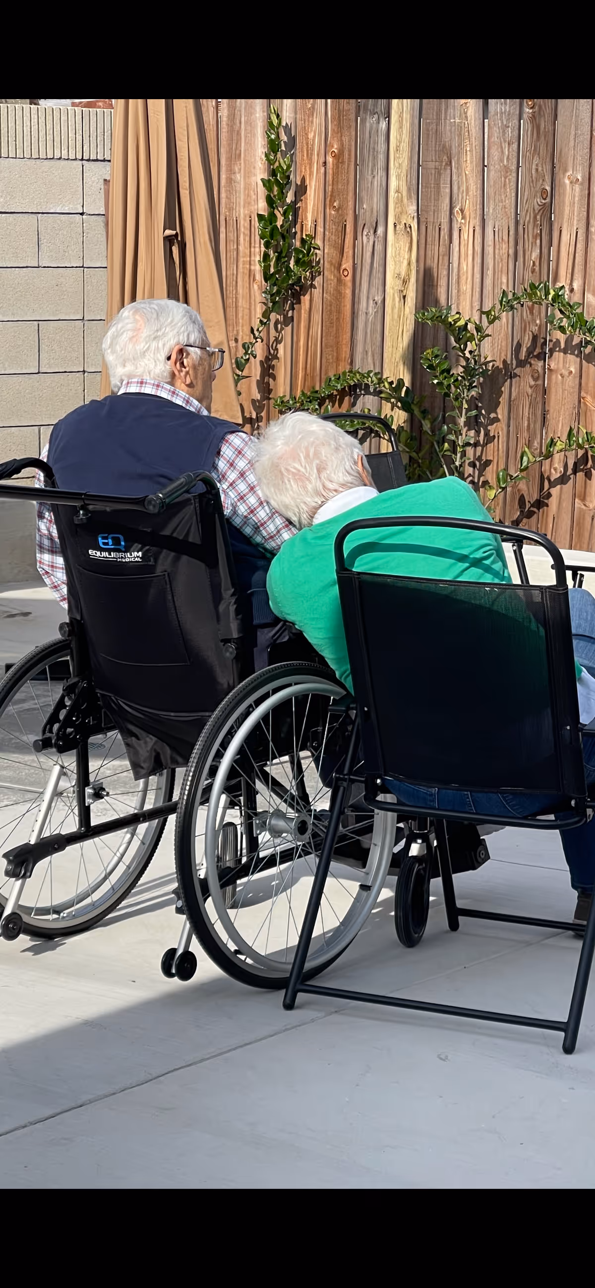 Two elderly people in wheelchairs sit outside near a wooden fence.