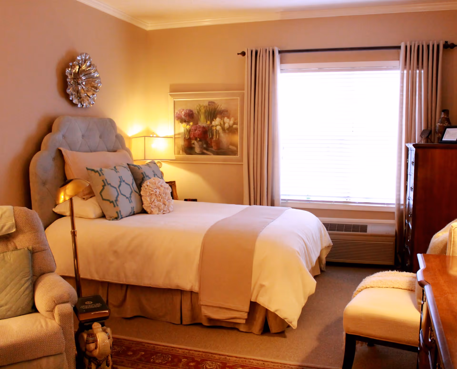 A cozy bedroom with a neatly made bed featuring multiple decorative pillows and a beige throw blanket. There is a cushioned armchair with a pillow on the left side, a wooden dresser on the right, and a window with beige curtains letting in natural light. The walls are painted a warm beige color and decorated with a floral painting and a metallic wall ornament. A table lamp provides soft lighting beside the bed.