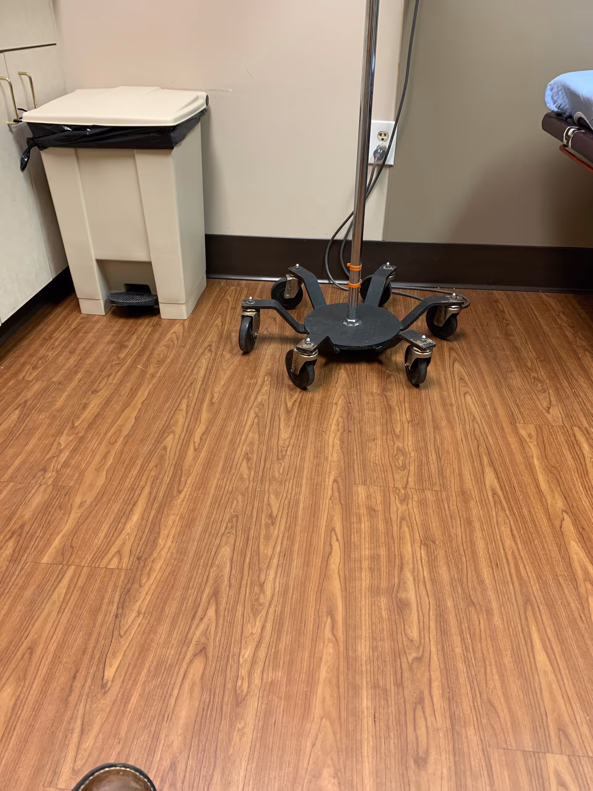 Hospital room corner with a wheeled IV stand, trash can, and wood-look floor.